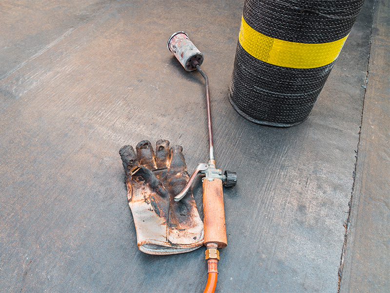 Werkzeuge und Material für die Abdichtung – Bitumenbahn, Propan-Flamme und Schutzhandschuhe für eine fachgerechte Balkonsanierung Abdichtung.